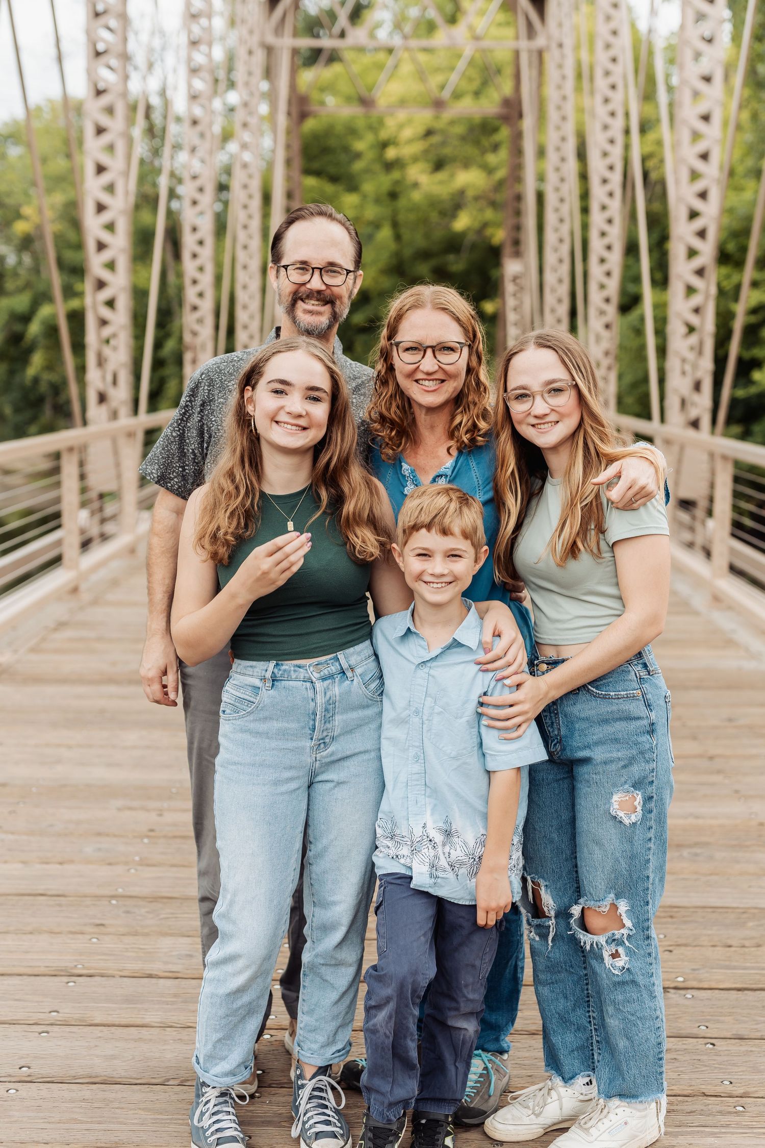 A happy family of five poses together on a wooden bridge surrounded by greenery on a sunny day.