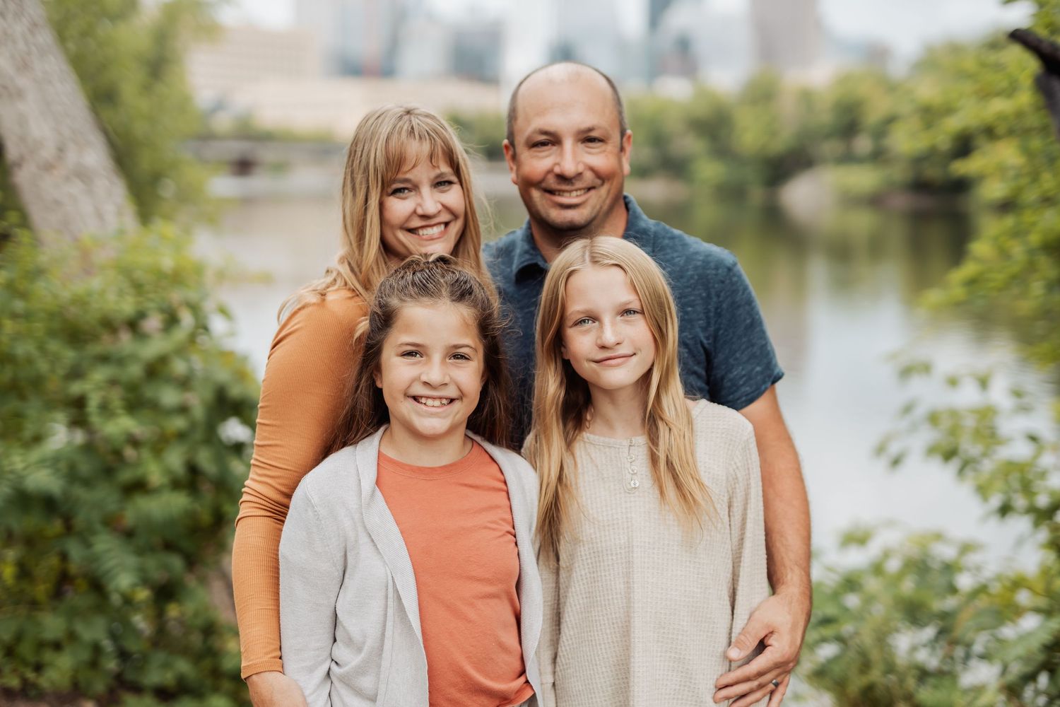 A happy family of four poses together on a path beside a river on a sunny autumn day.