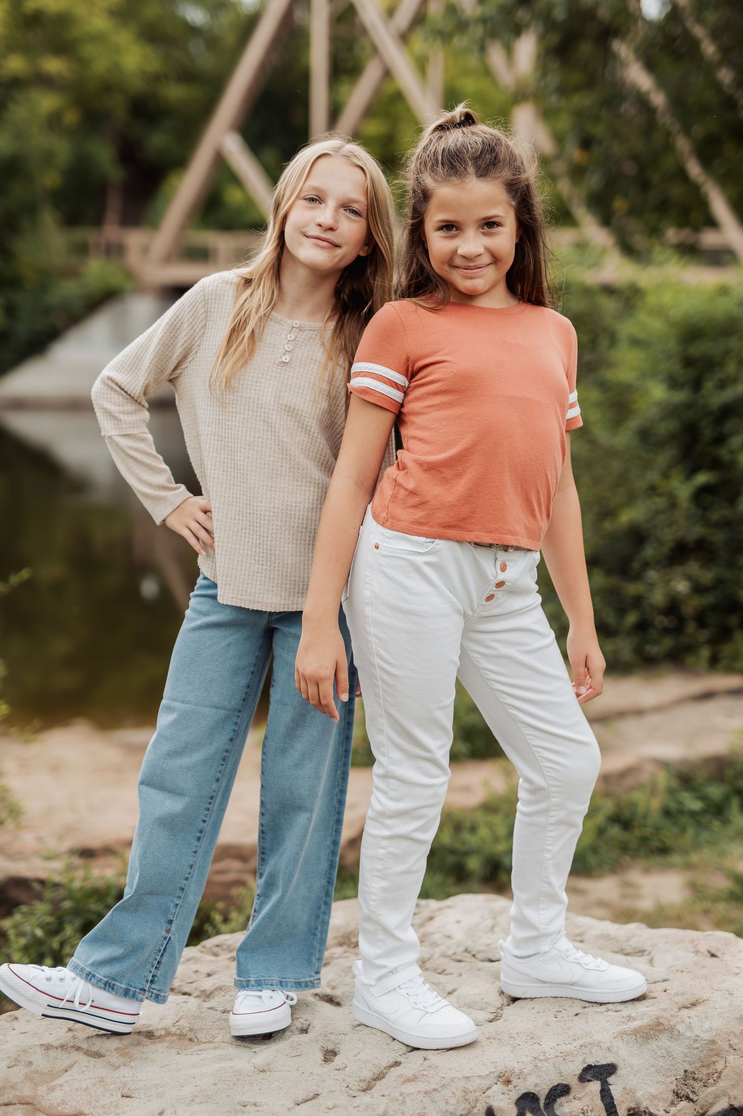 Two children posing outdoors near a pond wearing casual outfits with a cream sweater and orange t-shirt.