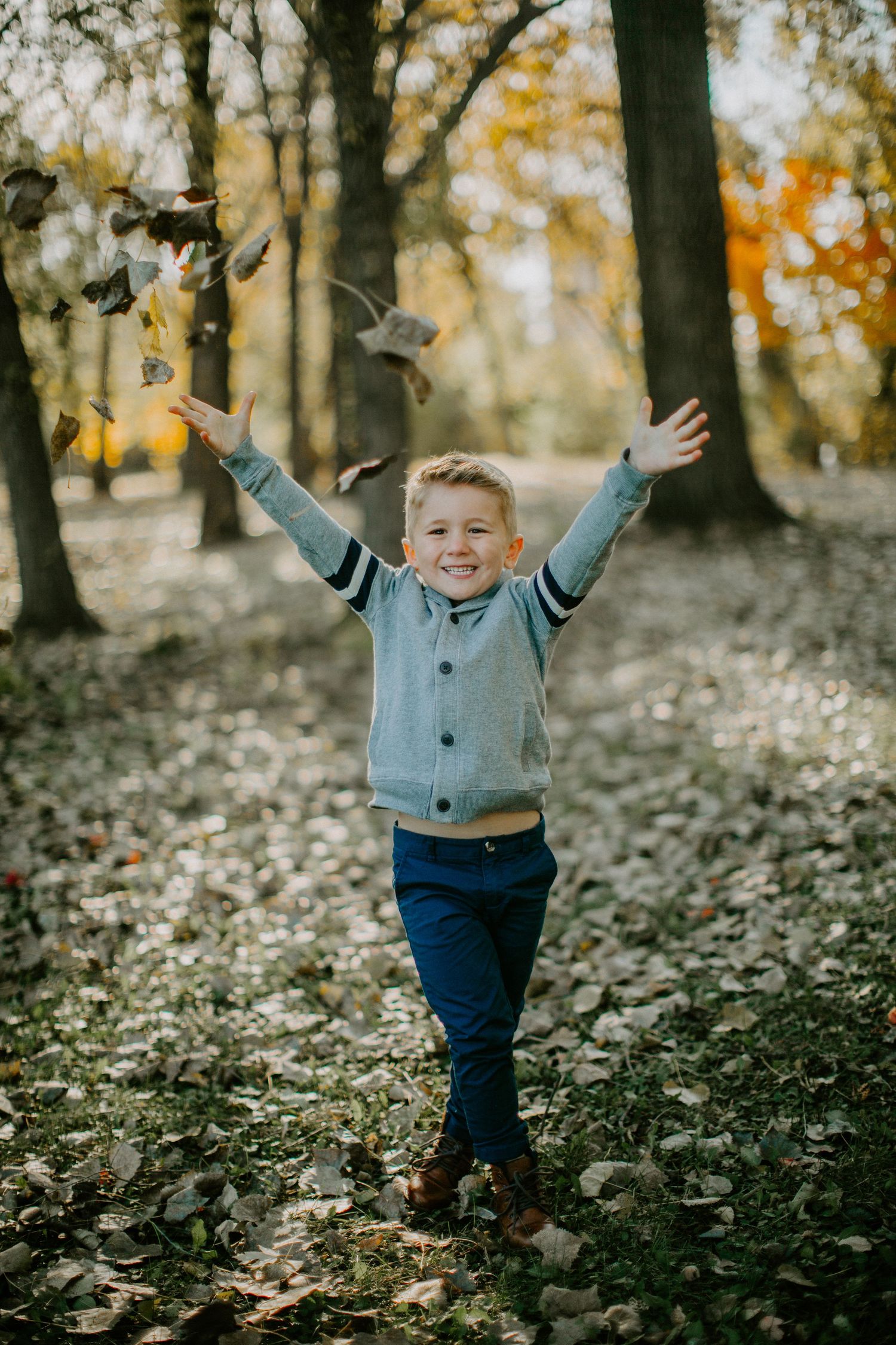 Joyful child throws autumn leaves in the air while wearing a gray cardigan and jeans in a wooded park setting.