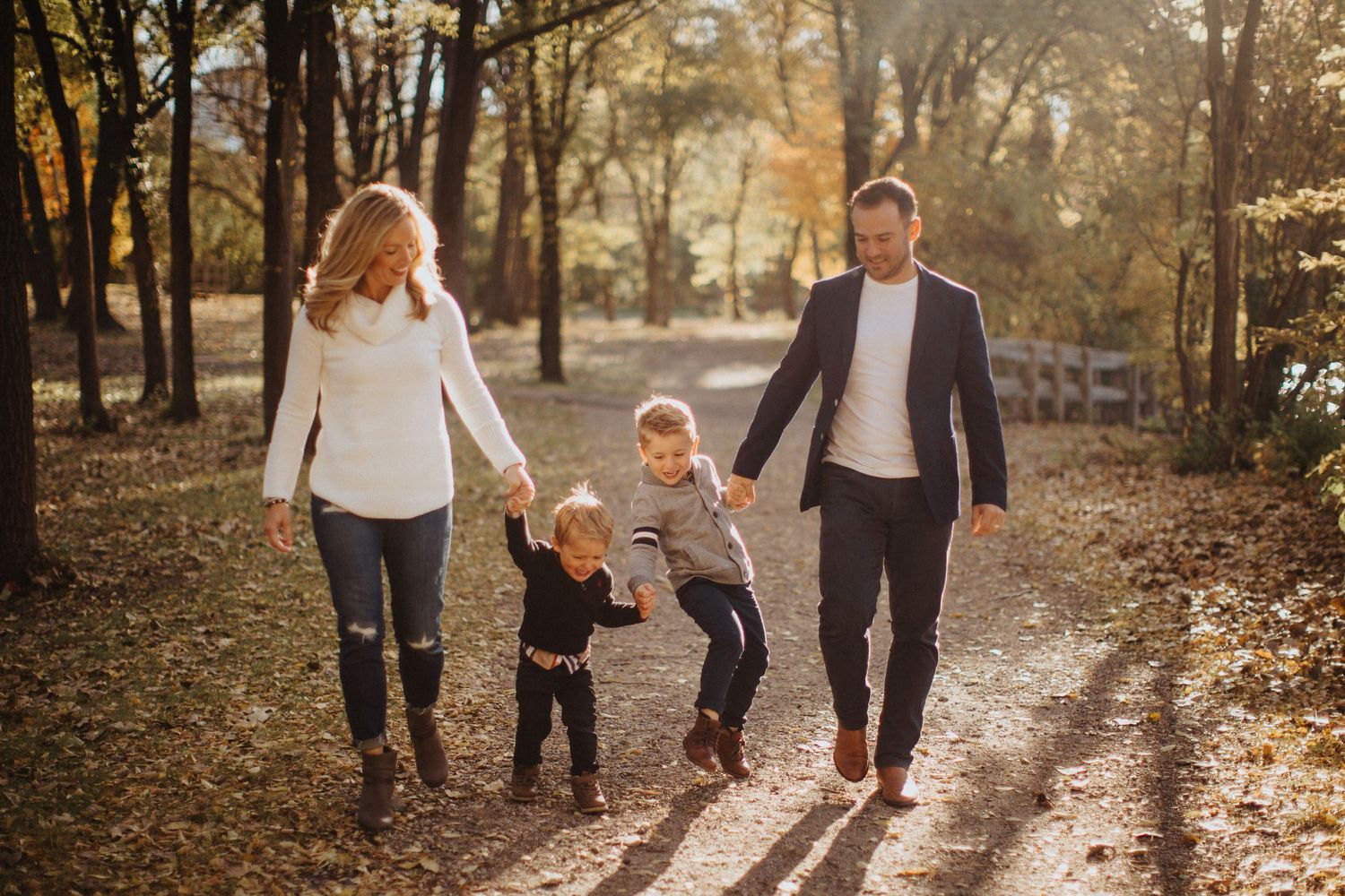 Family walks together holding hands on a sunlit autumn forest path surrounded by golden fallen leaves.