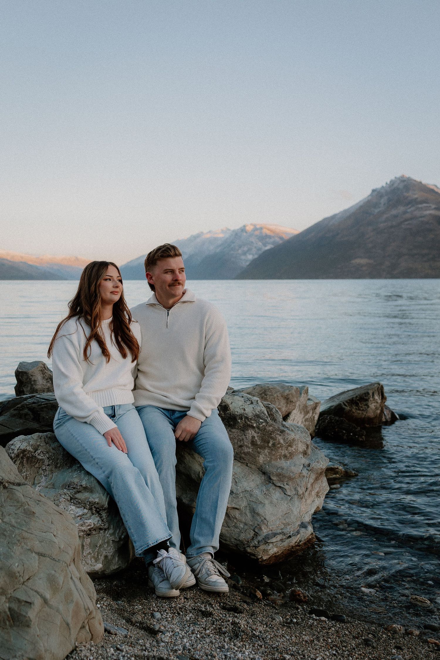 Two people walking through golden grass field with mountains in background during sunset.