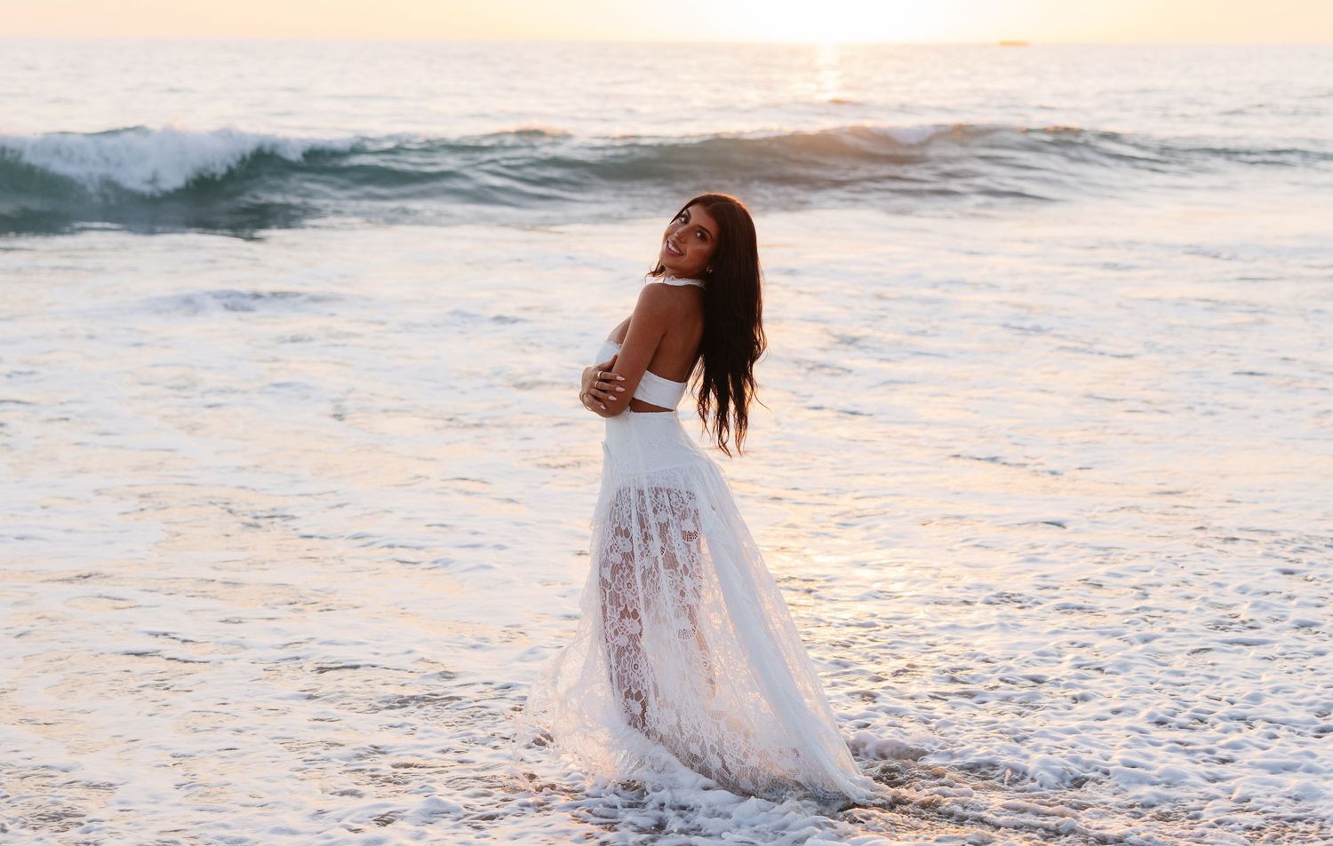 Woman in white lace dress standing by ocean waves at sunset.