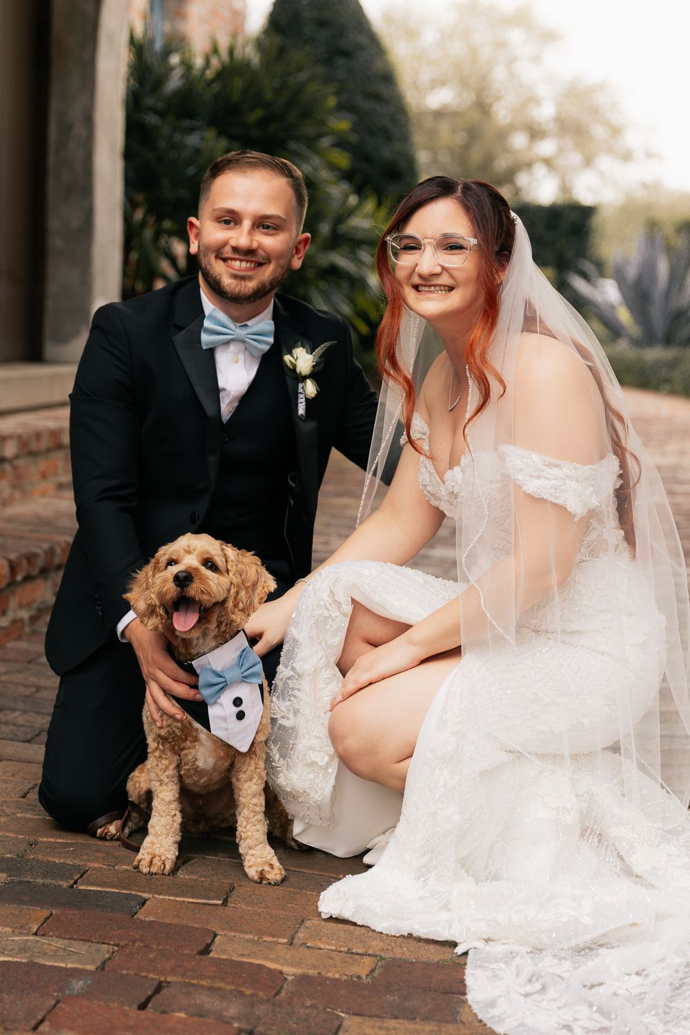 Smiling bride in a lace gown and veil crouches beside her groom in a blue bow tie and their fluffy dog in a tuxedo bandana.
