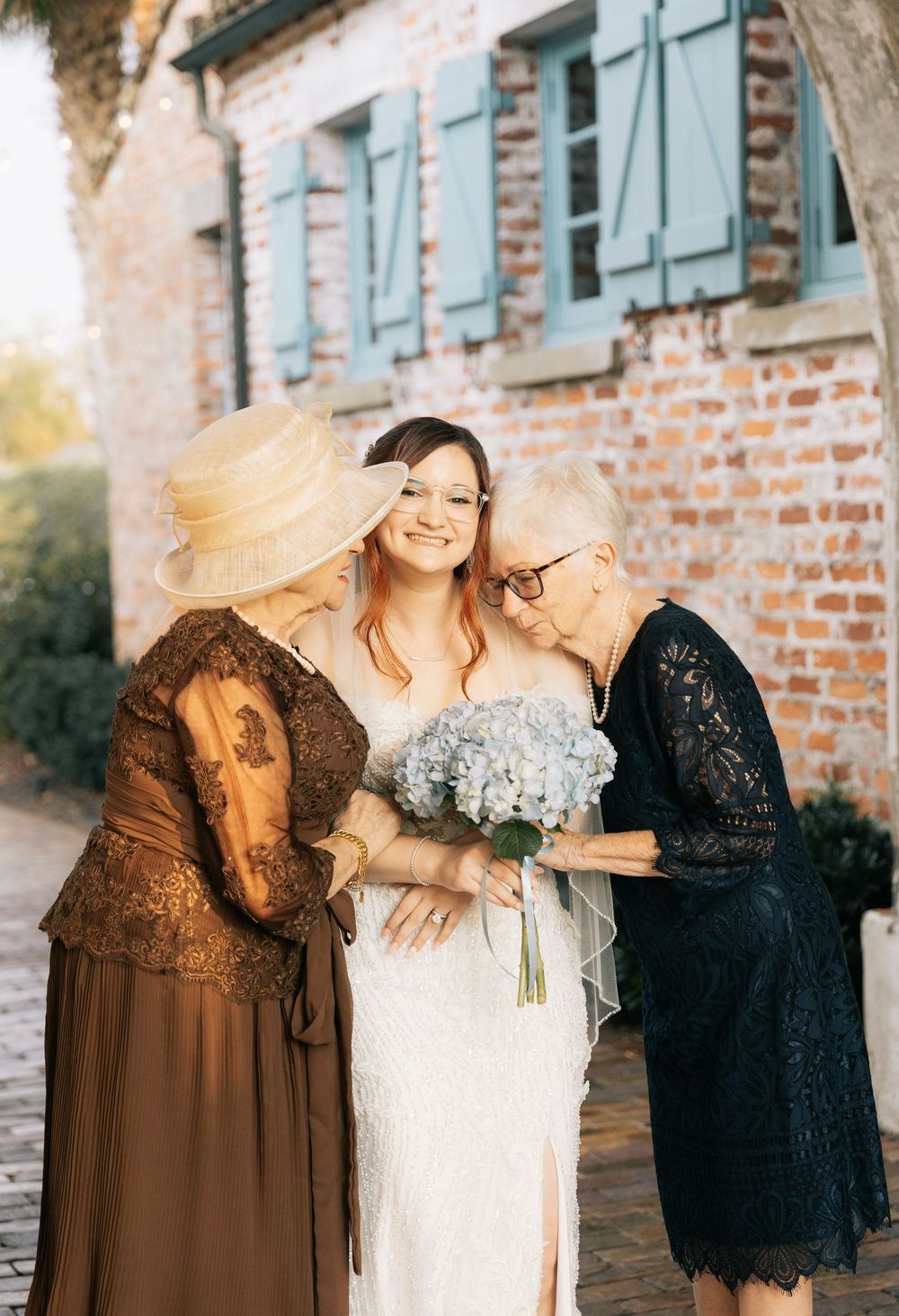 Smiling bride holding a blue hydrangea bouquet is embraced by two older women in front of a rustic brick building.