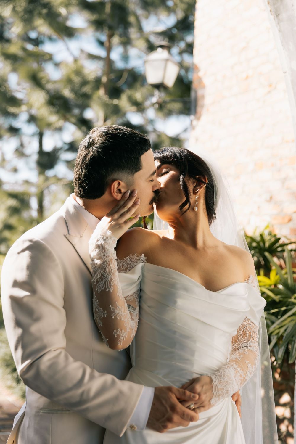Groom in a cream suit kisses his bride, who wears a lace-sleeved white gown and a veil, in a sunlit garden.