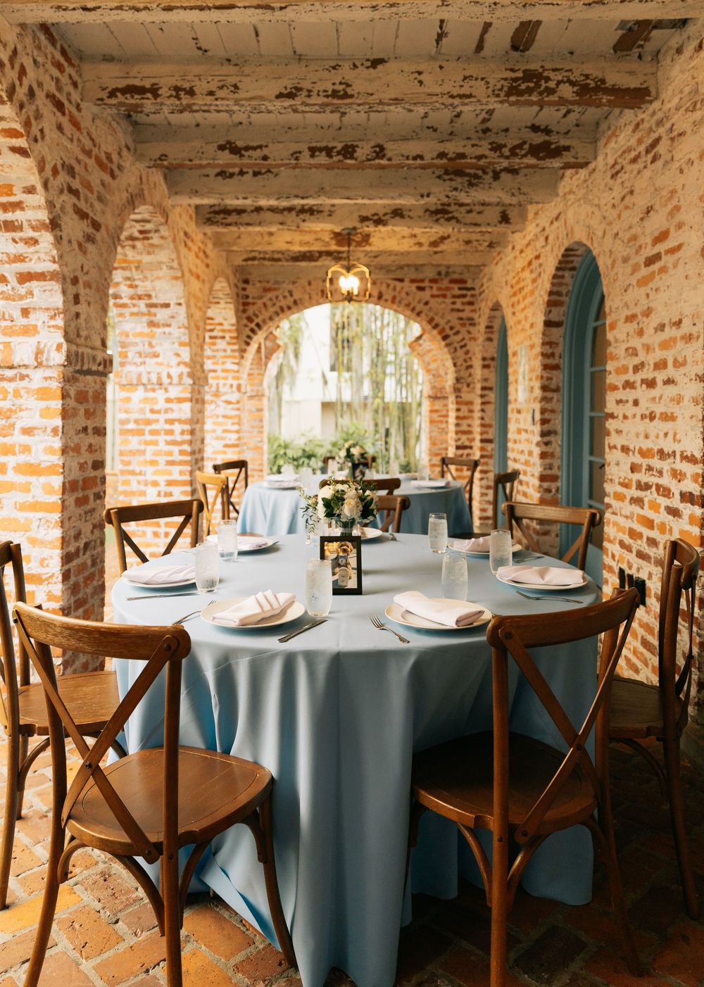 Round wedding reception table with a light blue linen and wooden chairs set beneath rustic brick arches.