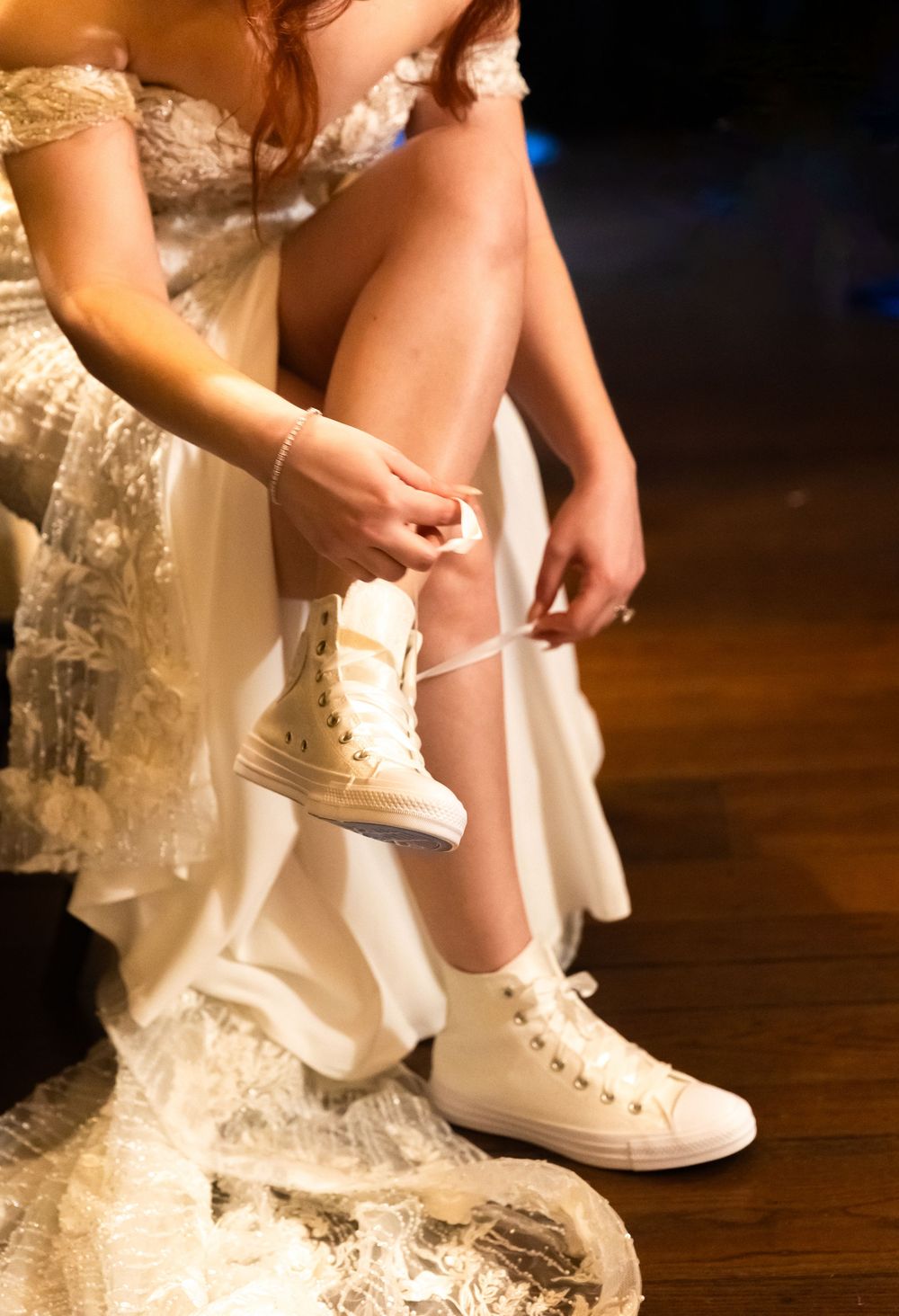 Bride in a lace wedding gown ties the laces of white high-top sneakers while seated on a wooden floor.