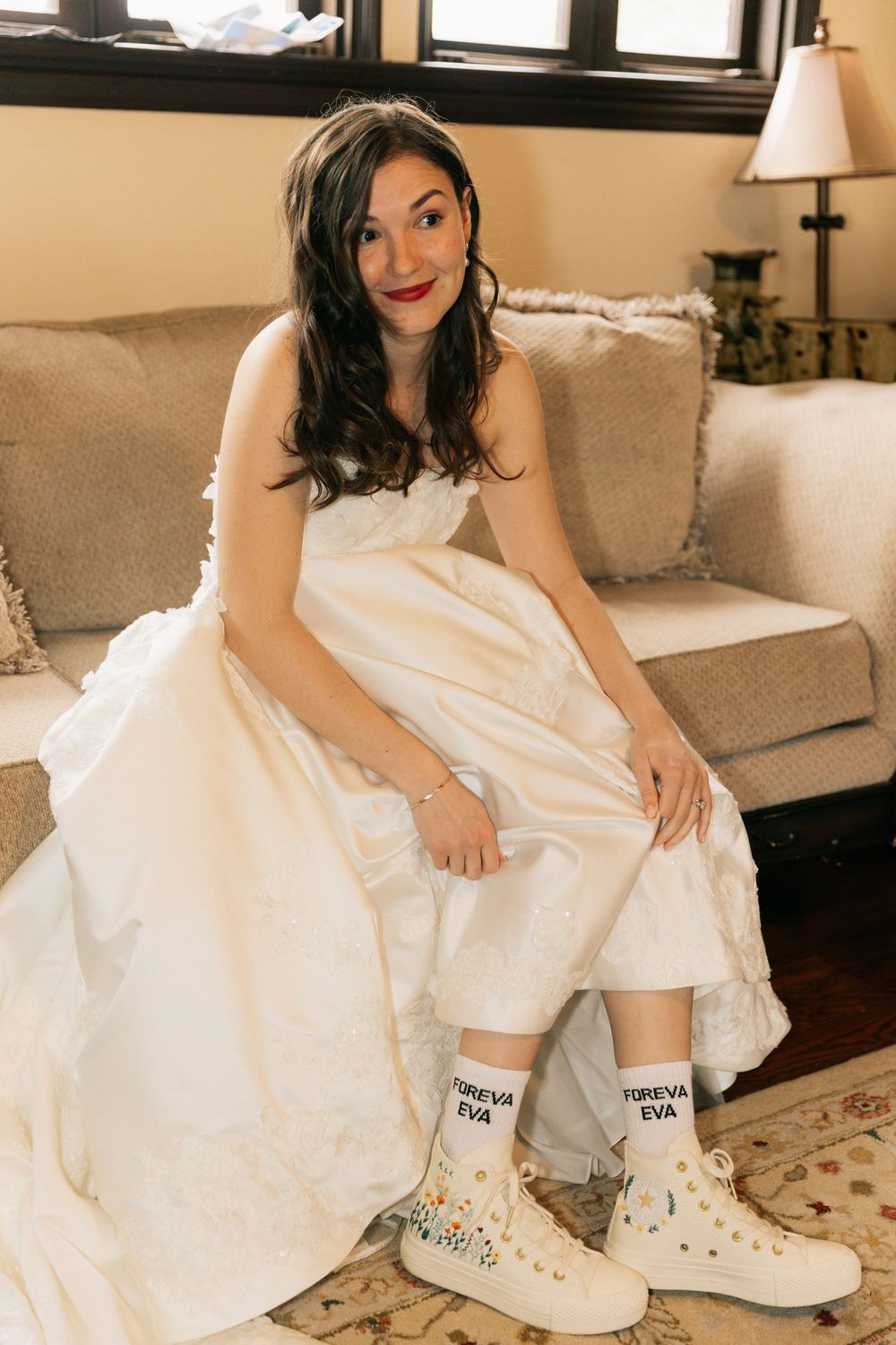 Smiling bride in a satin wedding gown sits on a sofa wearing white high-top sneakers and Forever Eva socks.