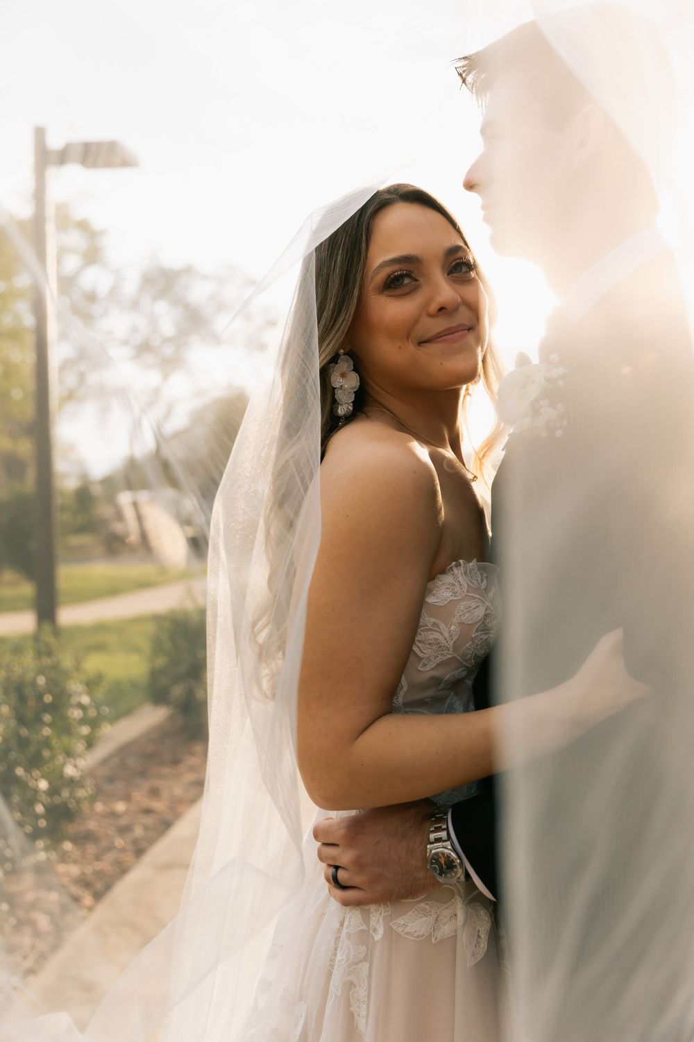 Smiling bride in a lace gown and flowing veil is held close by her groom in soft golden sunlight outdoors.