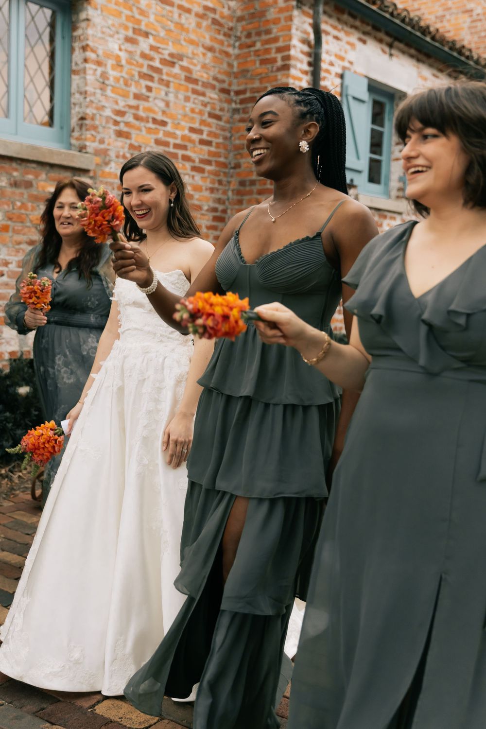 Bridesmaids in slate gray dresses laugh while holding orange bouquets next to a bride in a white gown by a brick wall.