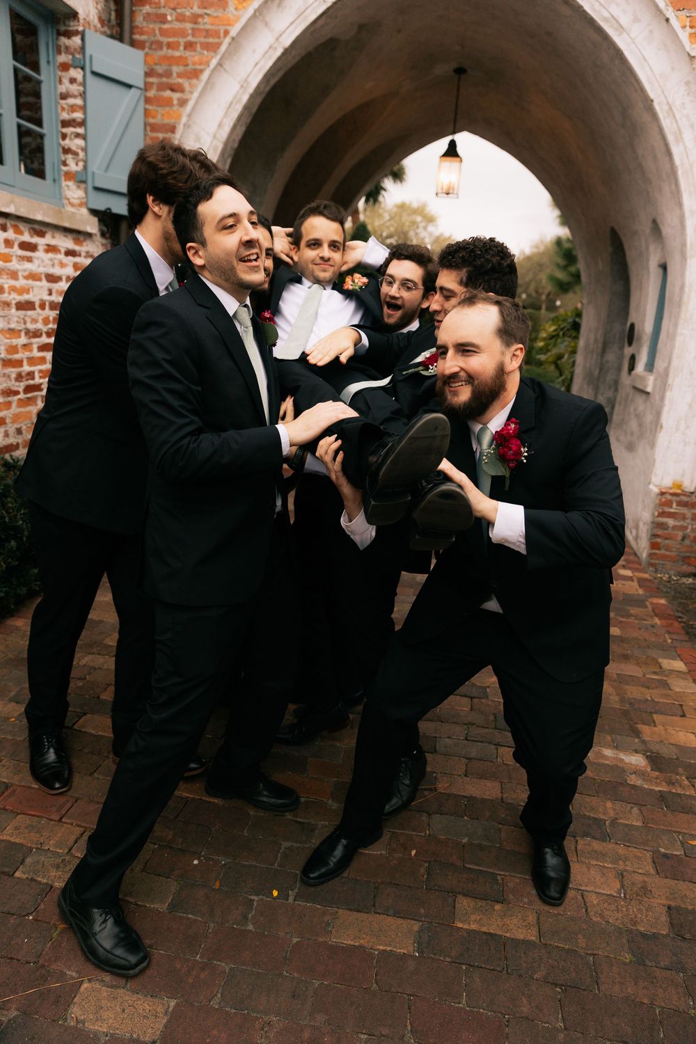 Laughing groomsmen in black suits playfully lift the groom into the air beneath a brick archway at a wedding venue.