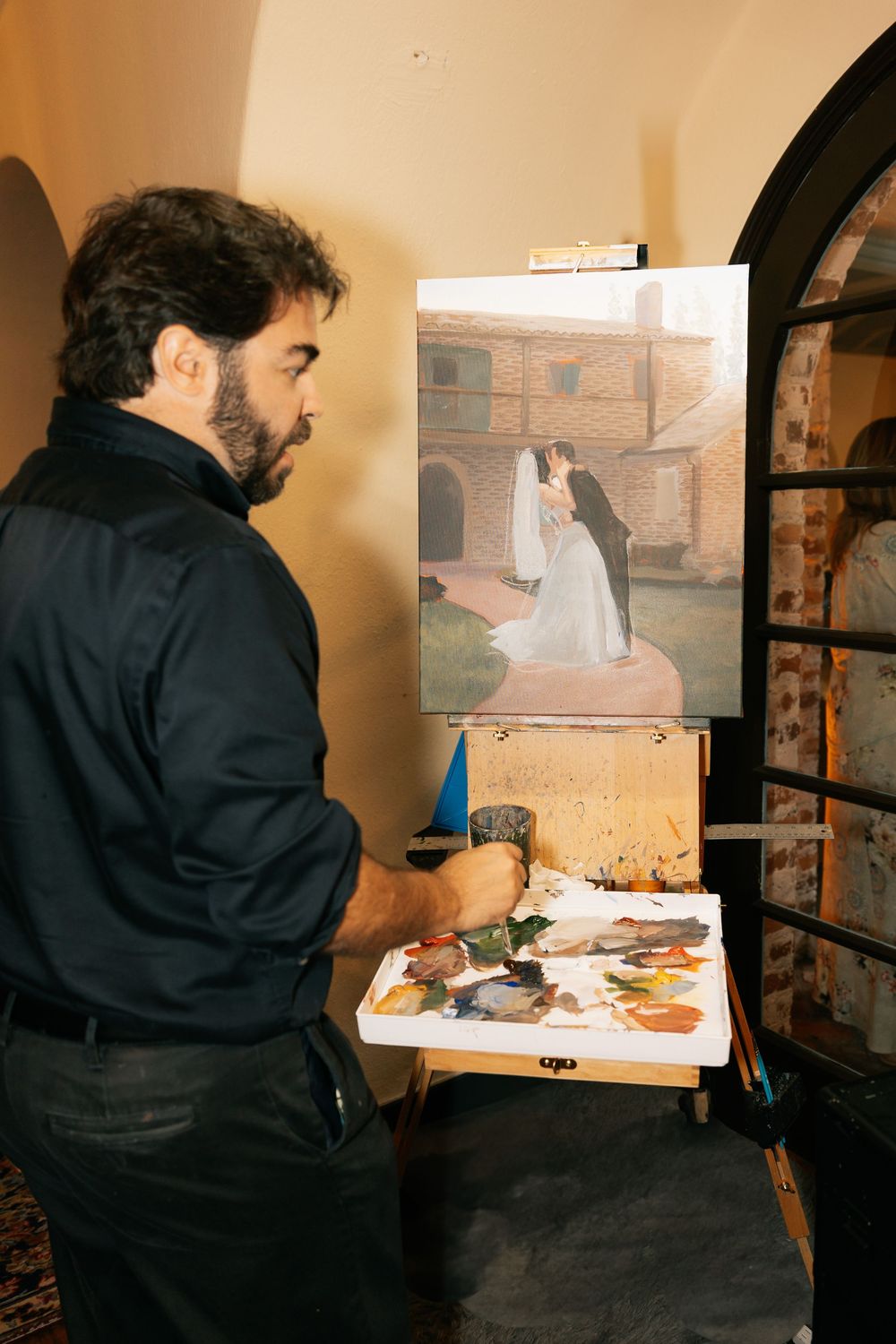 Live wedding painter works on an easel, creating an oil painting of a bride and groom in front of a brick building.