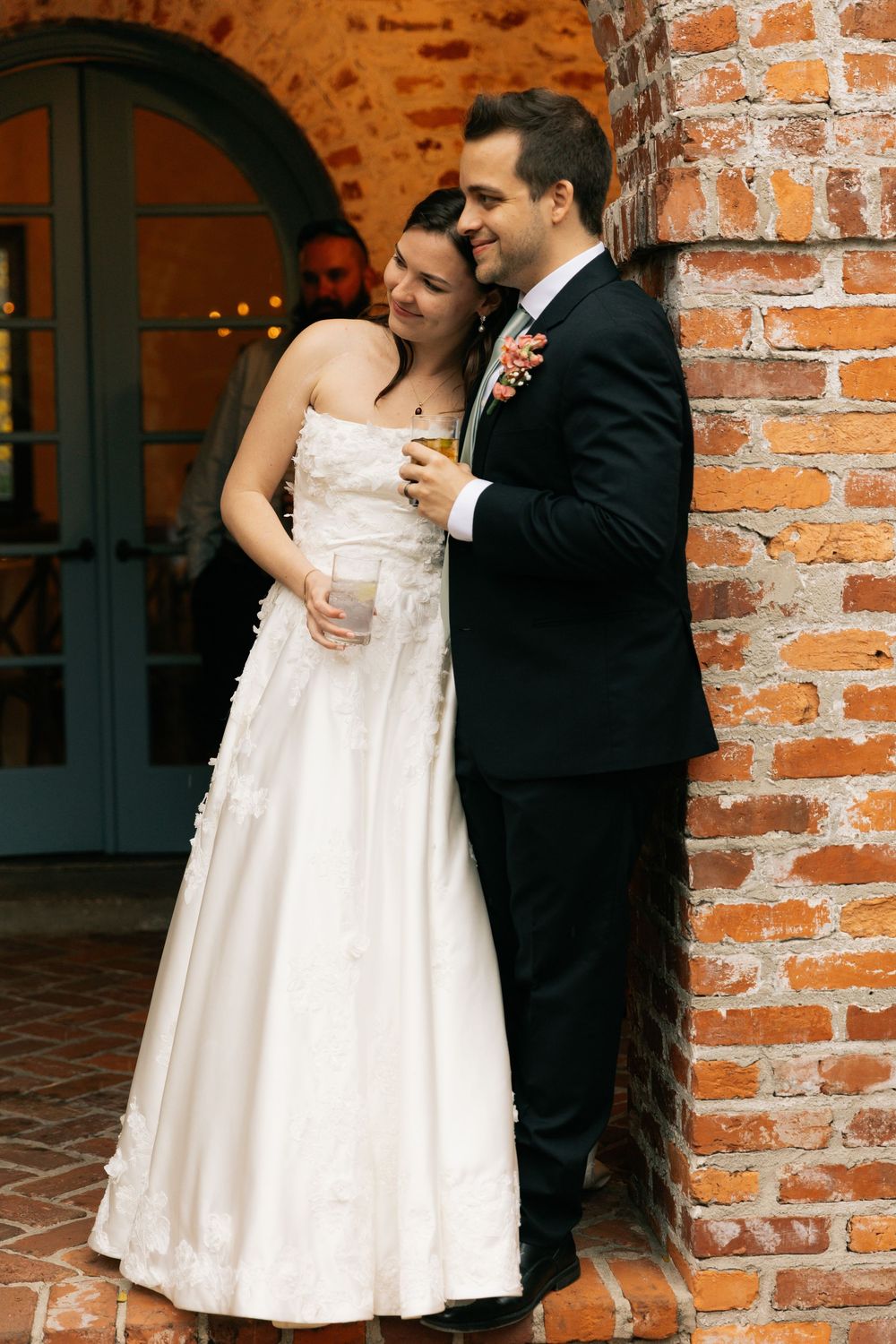 Bride in a strapless white gown and groom in a black suit with a floral boutonniere embrace against a brick wall holding drinks.