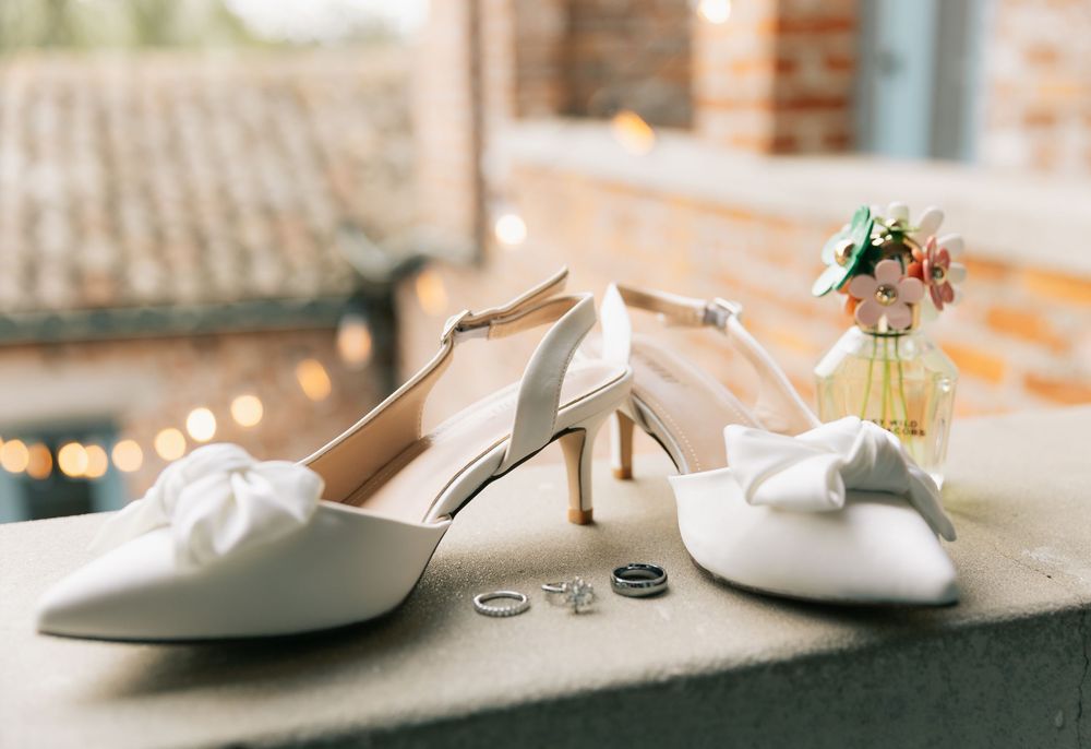 White bow-adorned wedding heels sit on a ledge beside two wedding rings and a small vase of flowers.