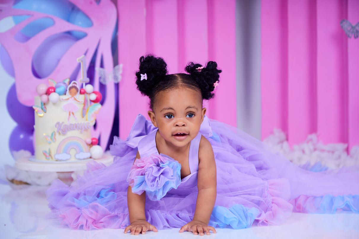 Child in purple tulle dress poses against pink backdrop with birthday cake.