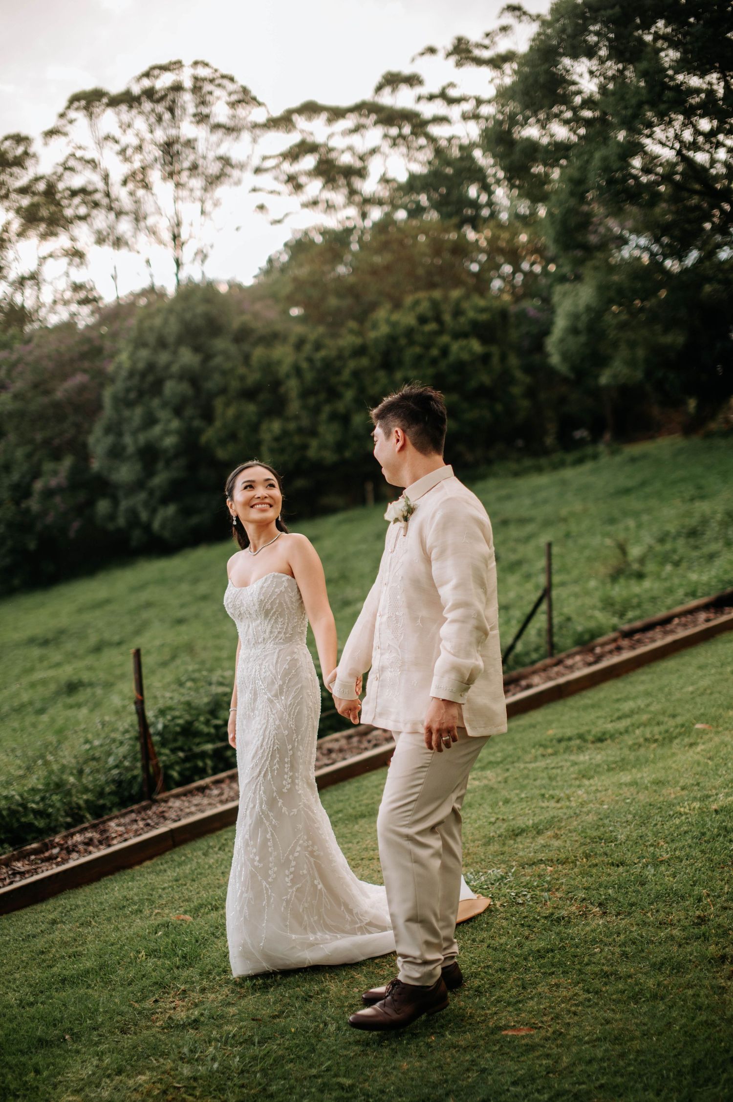 Bride in lace gown and groom in light suit walking on grassy hill with trees in background.