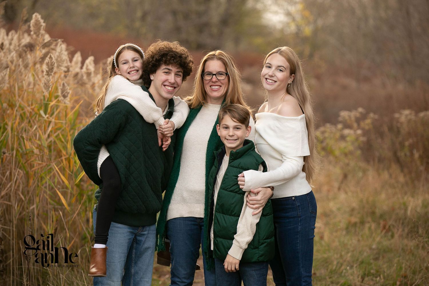 Family portrait of five people wearing coordinated white and dark green sweaters against an autumn background.