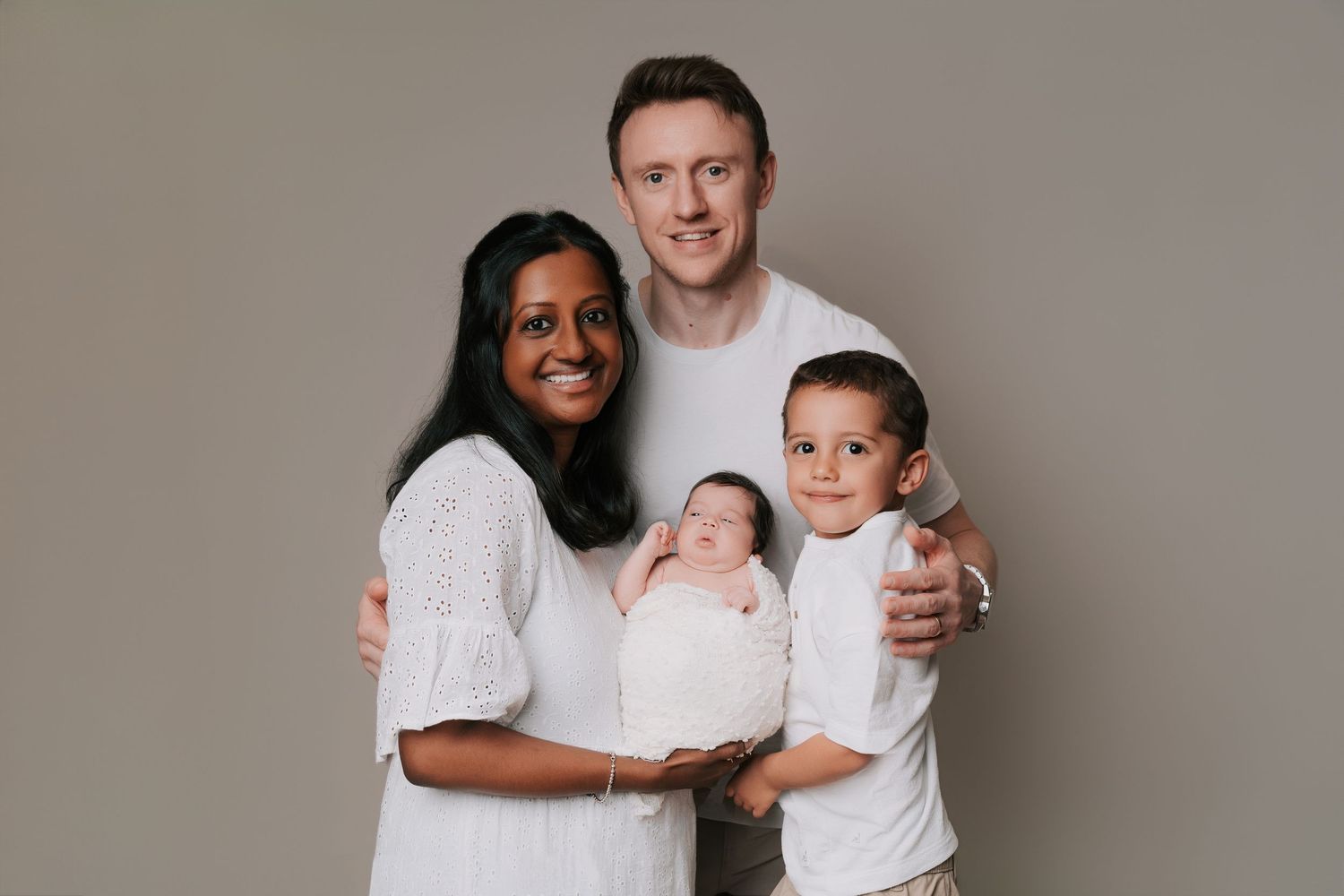 A family of four wearing white outfits poses together against a neutral gray background for a portrait.