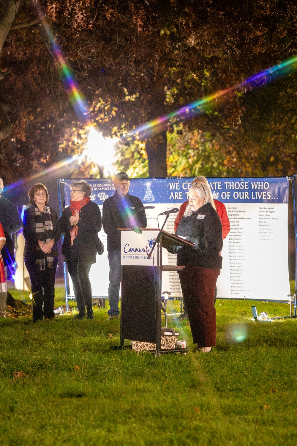 A small gathering takes place at dusk with a podium and banner display on a grassy area.