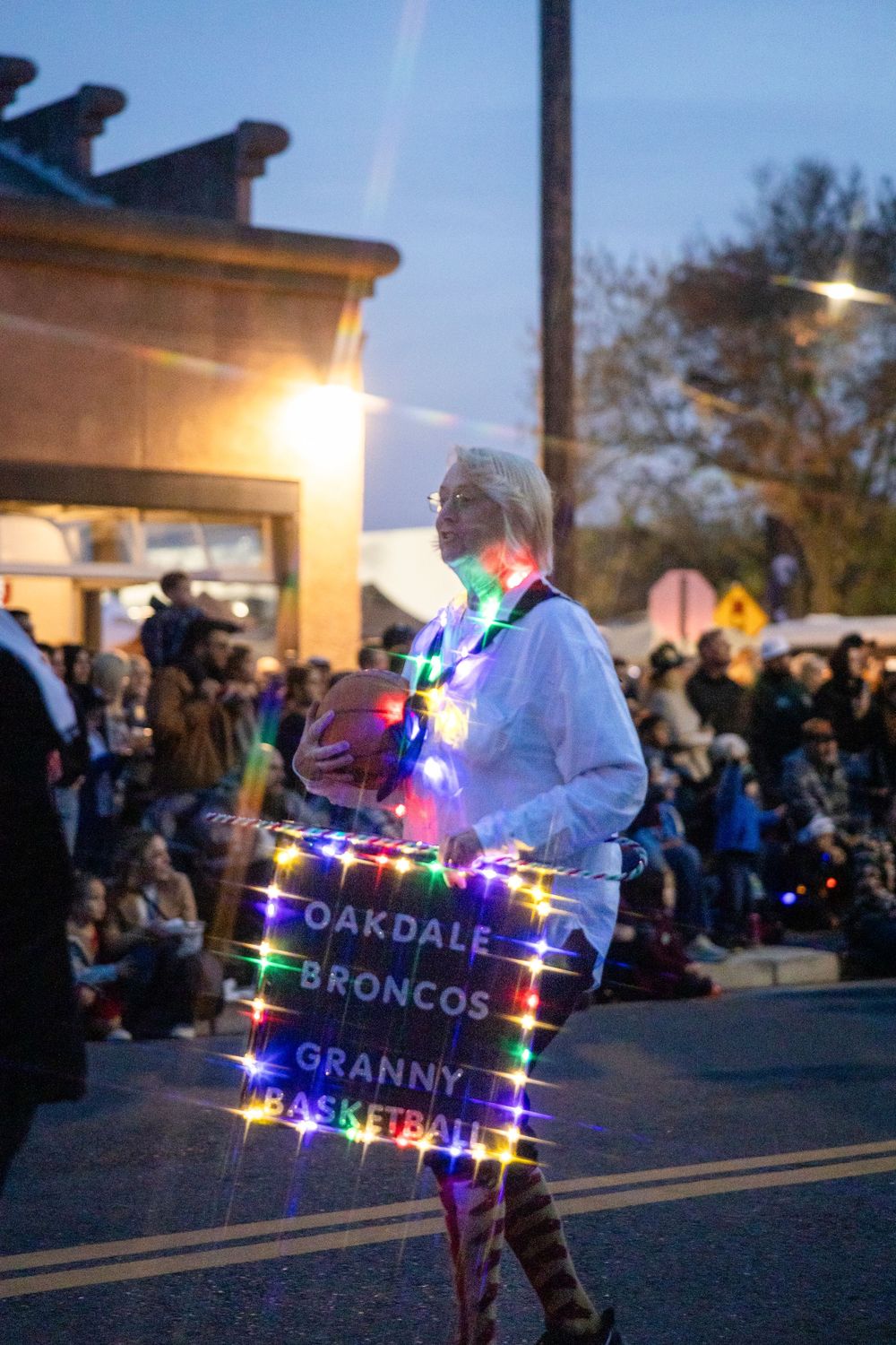 Person decorated with colorful Christmas lights holding a Oakdale Broncos basketball sign at a nighttime parade.