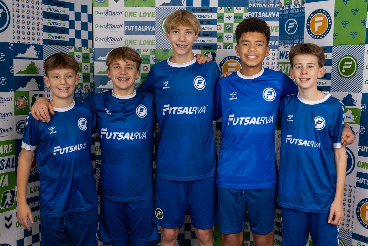 Youth futsal team members pose in matching blue jerseys against branded sponsorship wall.