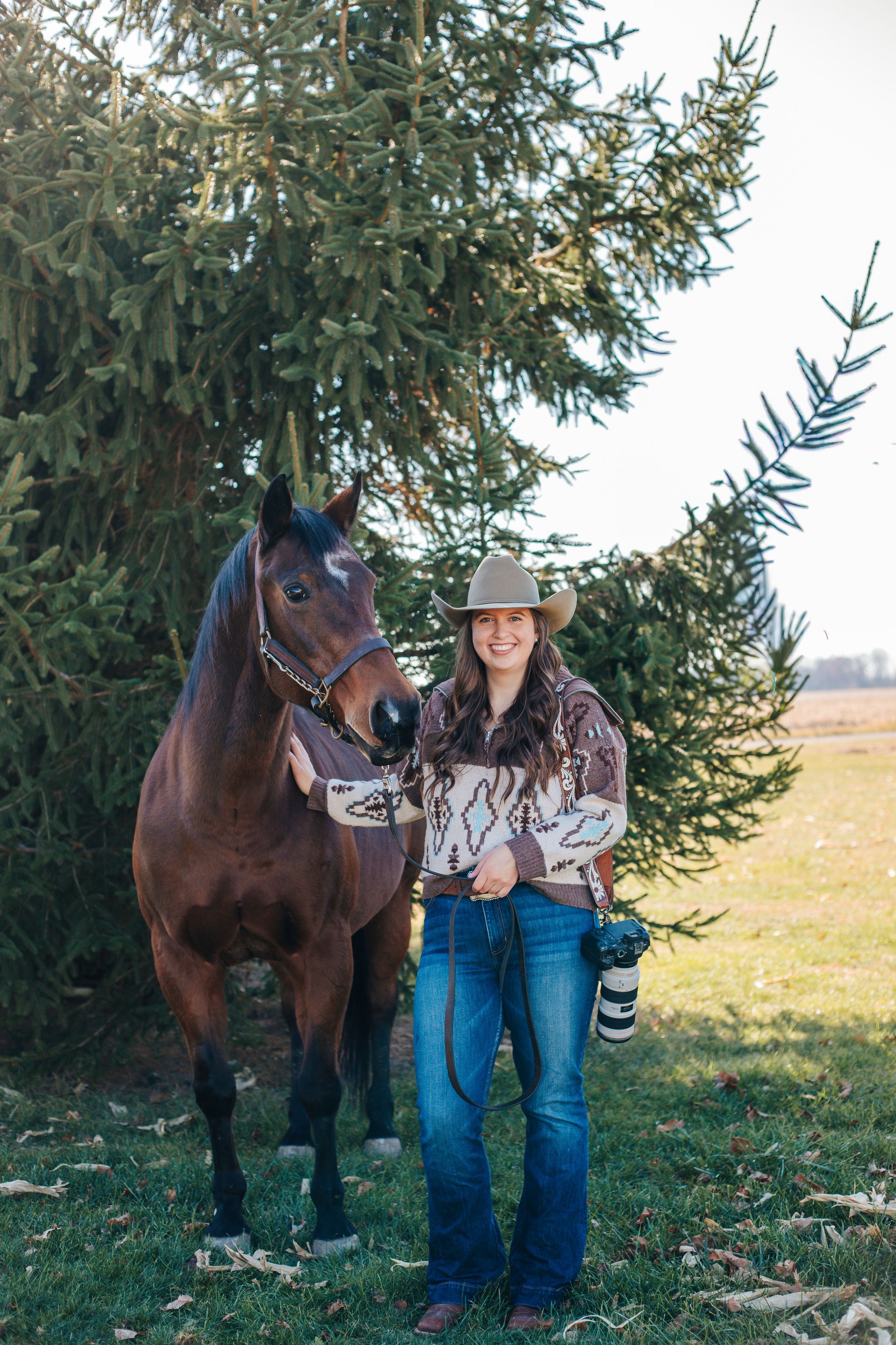 A bay horse stands beside someone in western wear and a cowboy hat near evergreen trees.