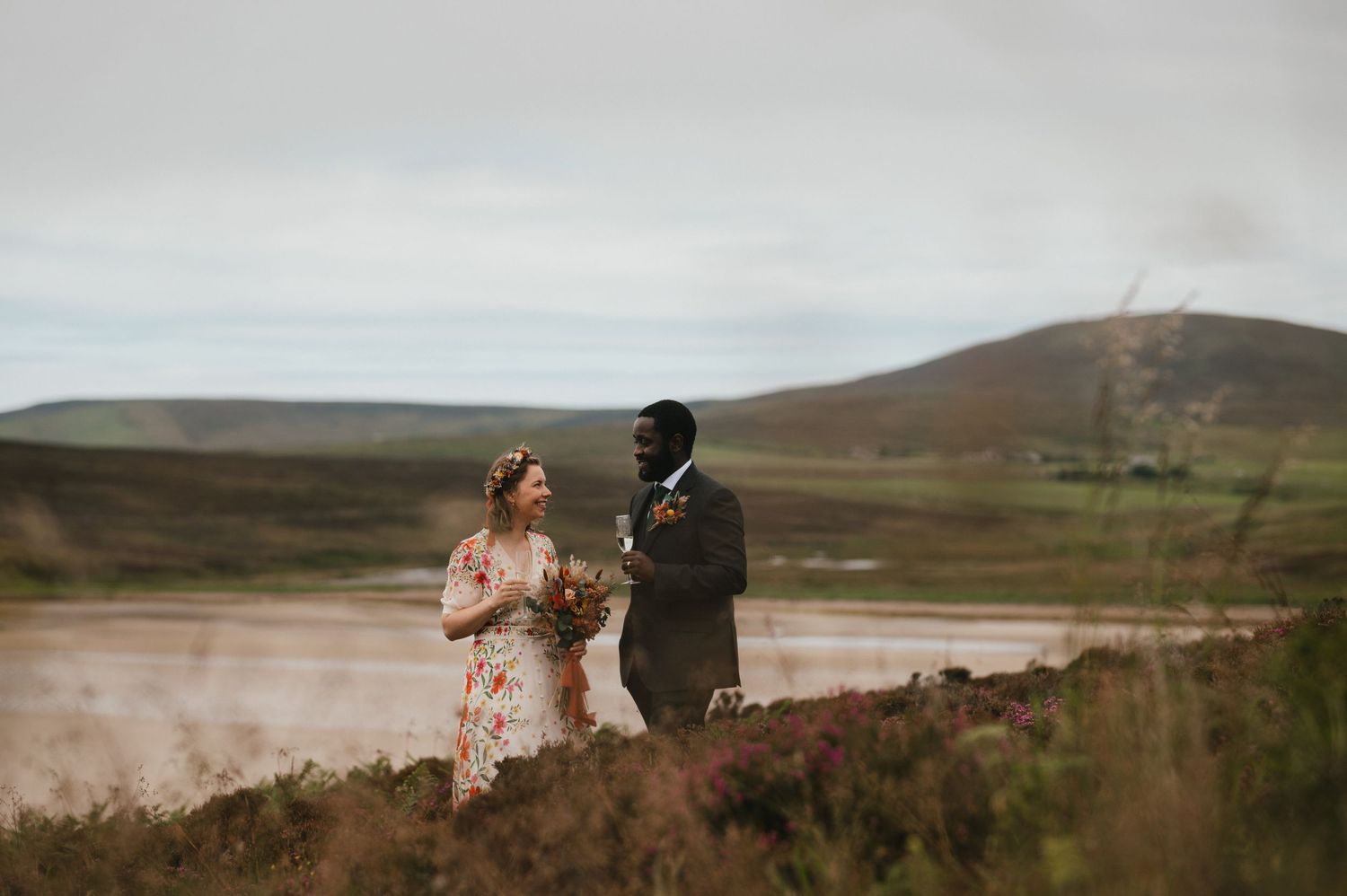 A romantic wedding couple stands on a hillside overlooking a misty Scottish landscape during golden hour.