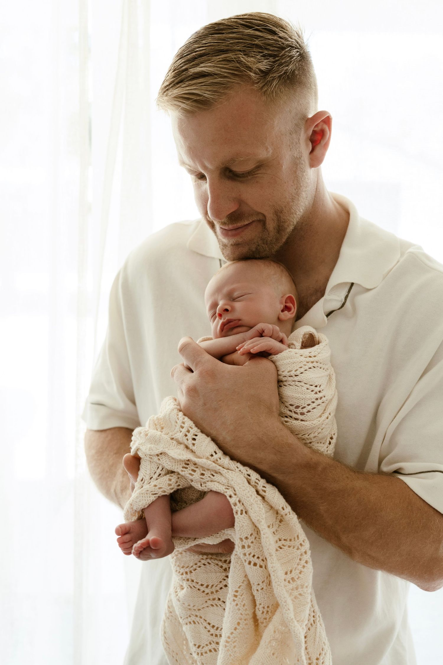 Proud father in white shirt gently cradling sleeping newborn wrapped in cream knit blanket by bright window.