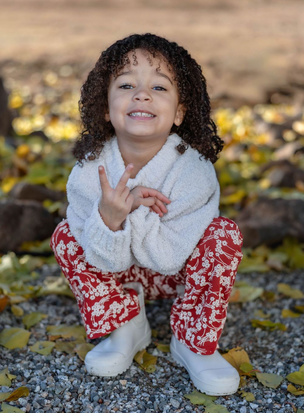 Child in festive red and white winter outfit poses on rocky ground with fallen leaves.