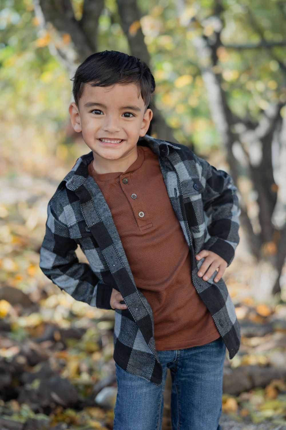 Child in plaid flannel shirt and jeans poses playfully outdoors during autumn.