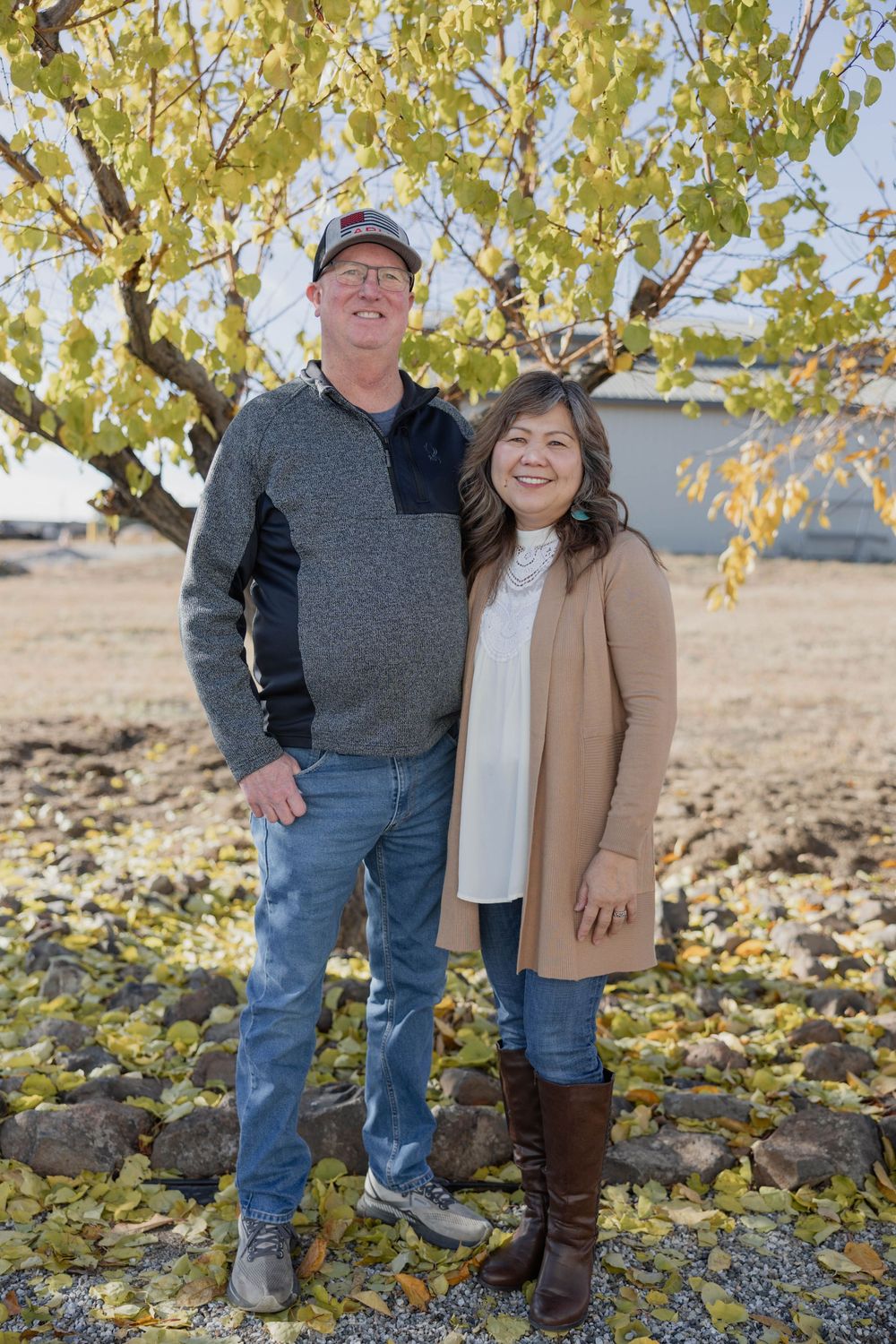 Couple poses together under yellow autumn trees in casual outdoor attire.
