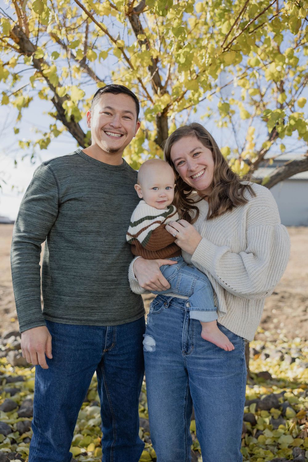Family of three poses together under yellow autumn trees for casual outdoor portrait.