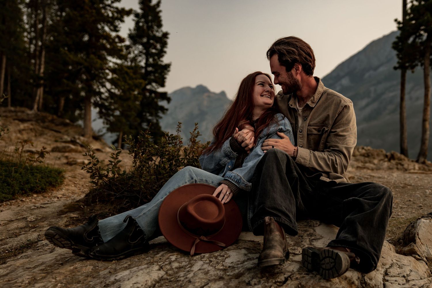 A couple sits together on a mountain ledge overlooking a scenic valley during sunset.