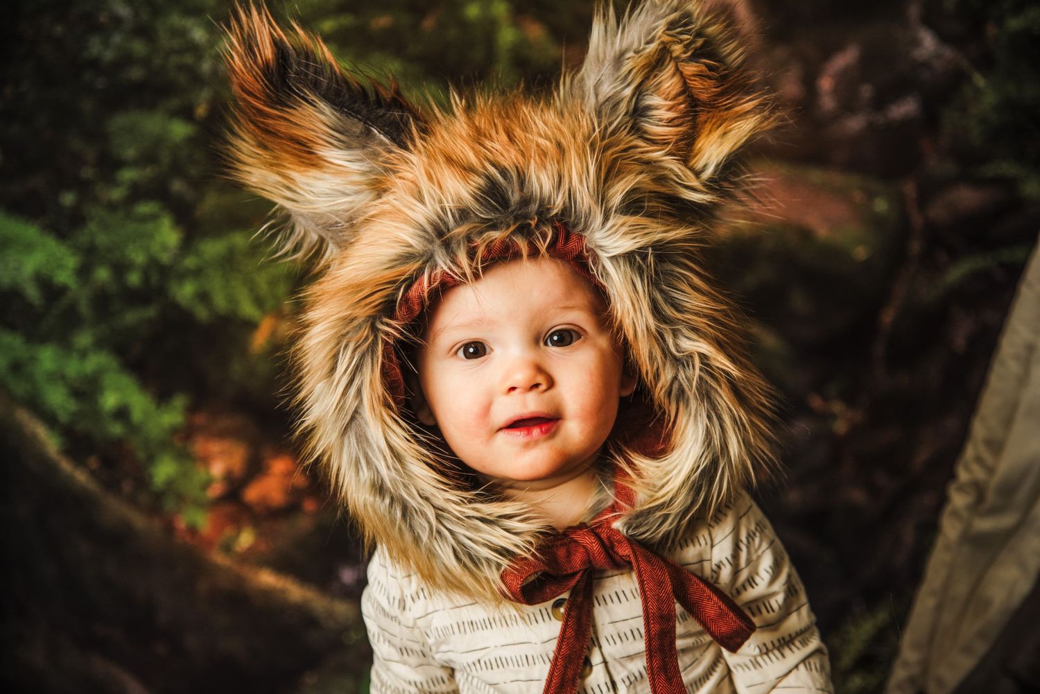 A young child wearing a furry fox-themed winter hood with pointed ears and a red bow in a woodland setting.