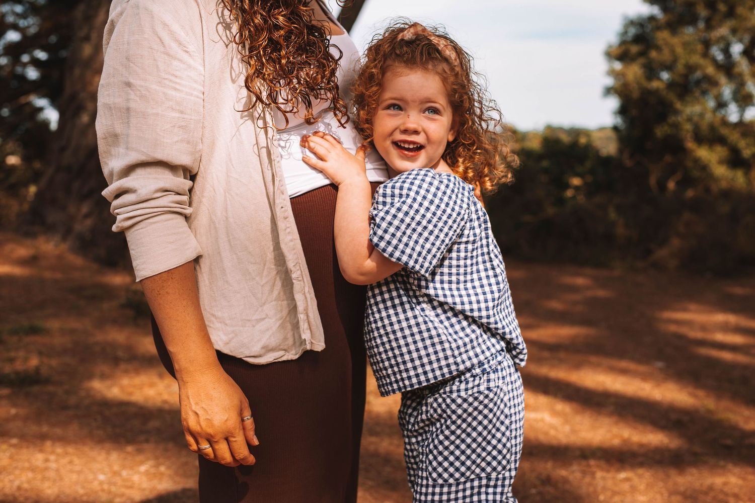A toddler in a gingham dress hugs their parent's leg during a warm sunset photoshoot in a wooded area.