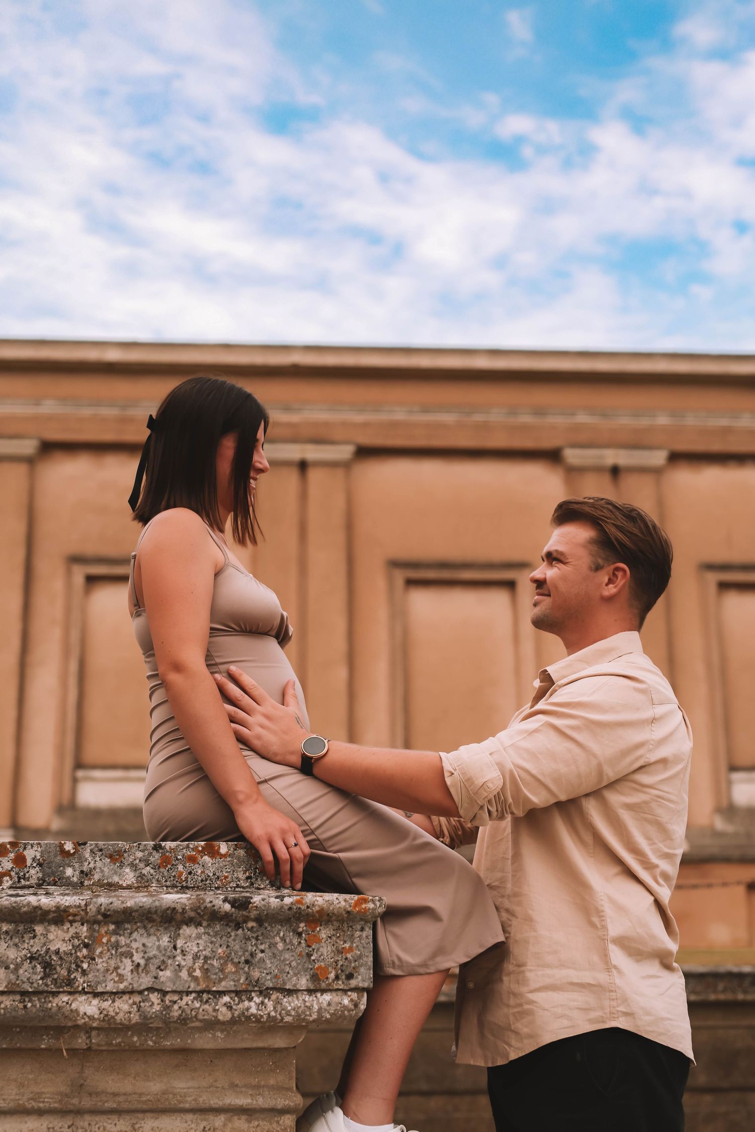 A couple shares a tender moment on stone steps against a peach-colored building under a bright blue sky.