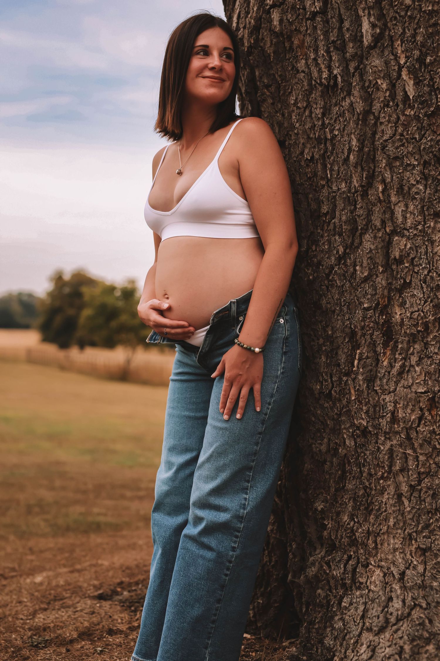 A pregnant woman in a white crop top and jeans leans against a tree trunk in a rural setting at sunset.