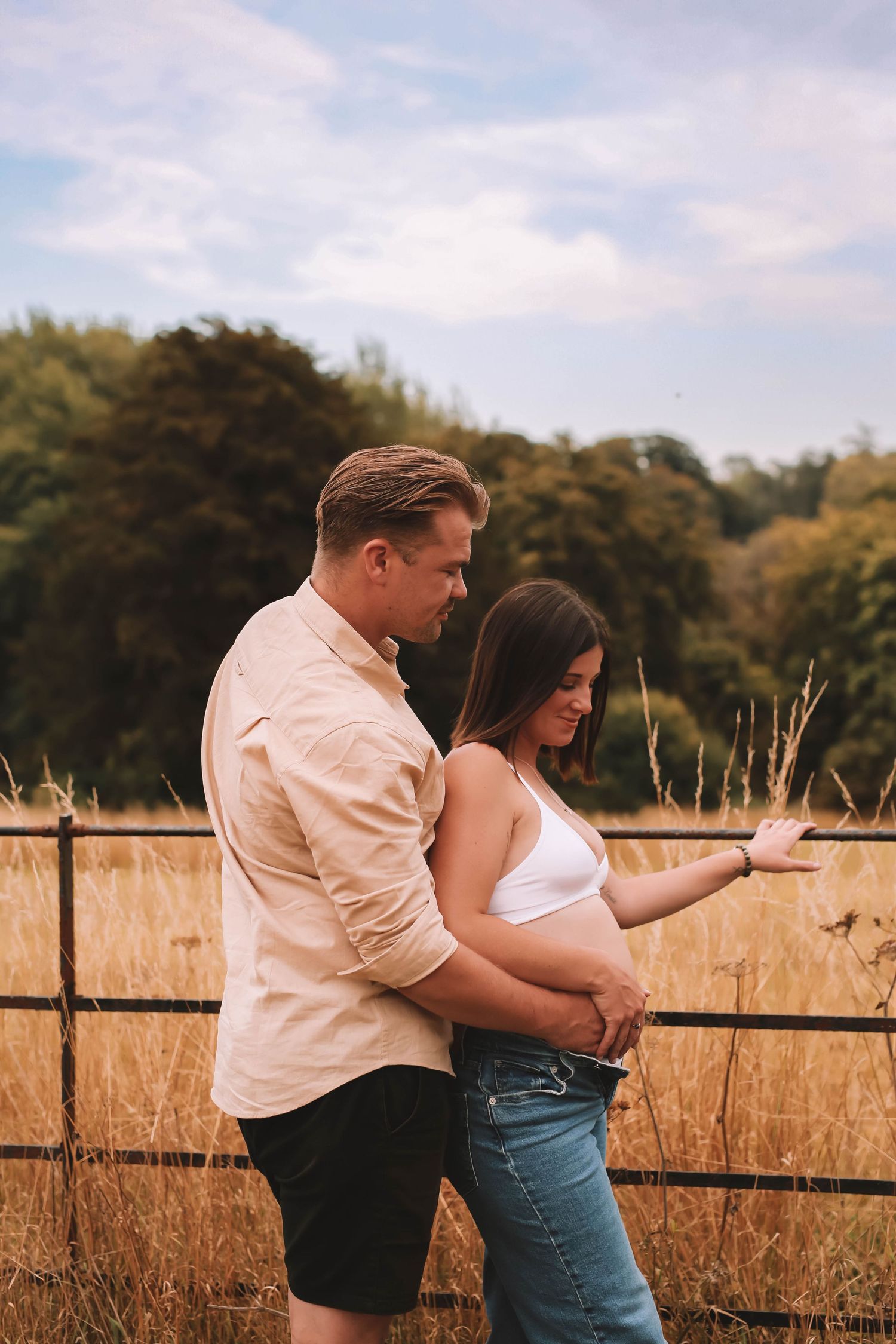 A couple embraces in a golden field at sunset with a rustic wooden fence in the background.