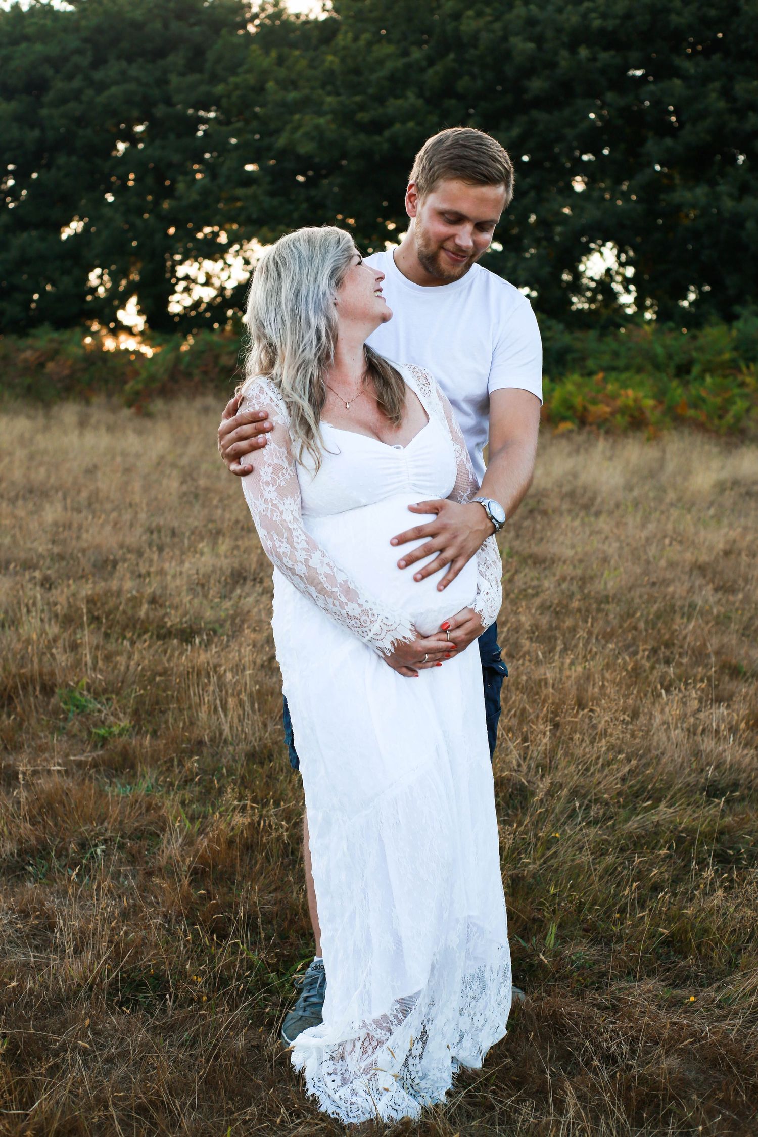 A couple shares a tender moment in a field at sunset, with the woman wearing a white lace maternity dress.
