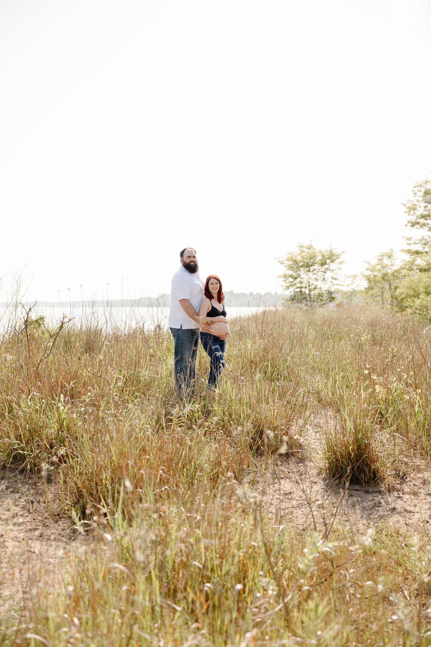 Couple embraces on a sandy beach dune surrounded by tall grass and coastal vegetation on a sunny day.