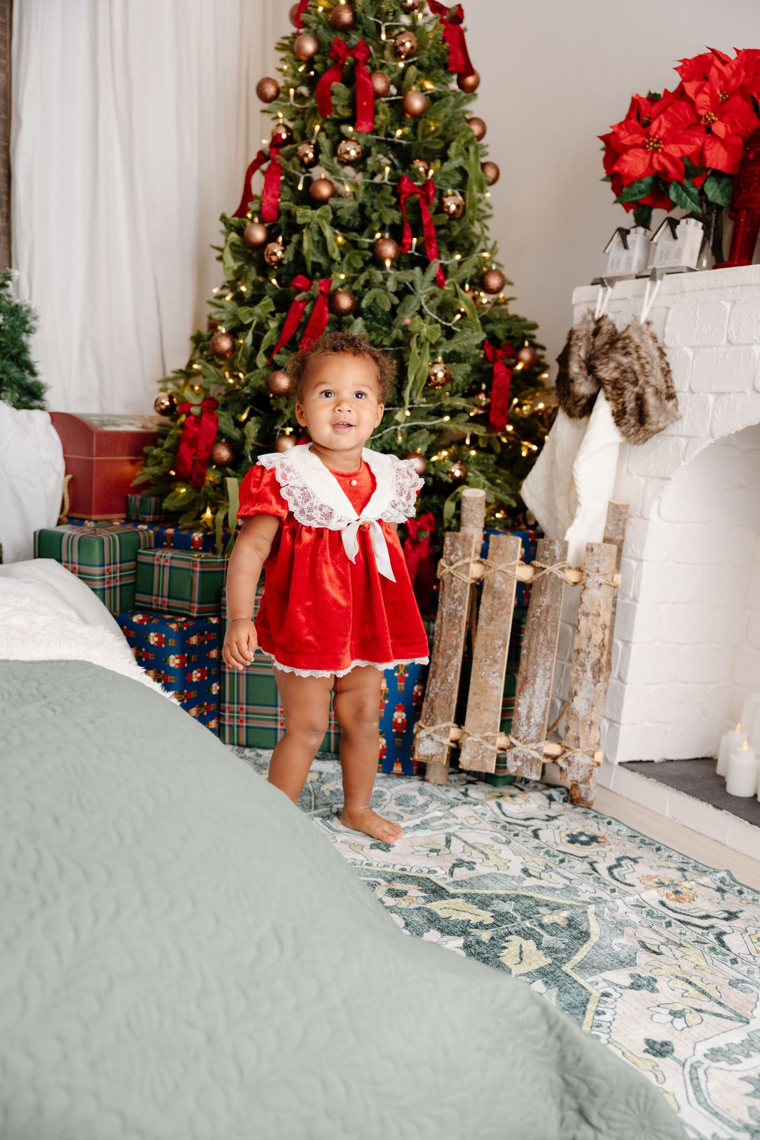A small child in a red holiday dress stands near decorated Christmas trees and wrapped presents in a festive room.