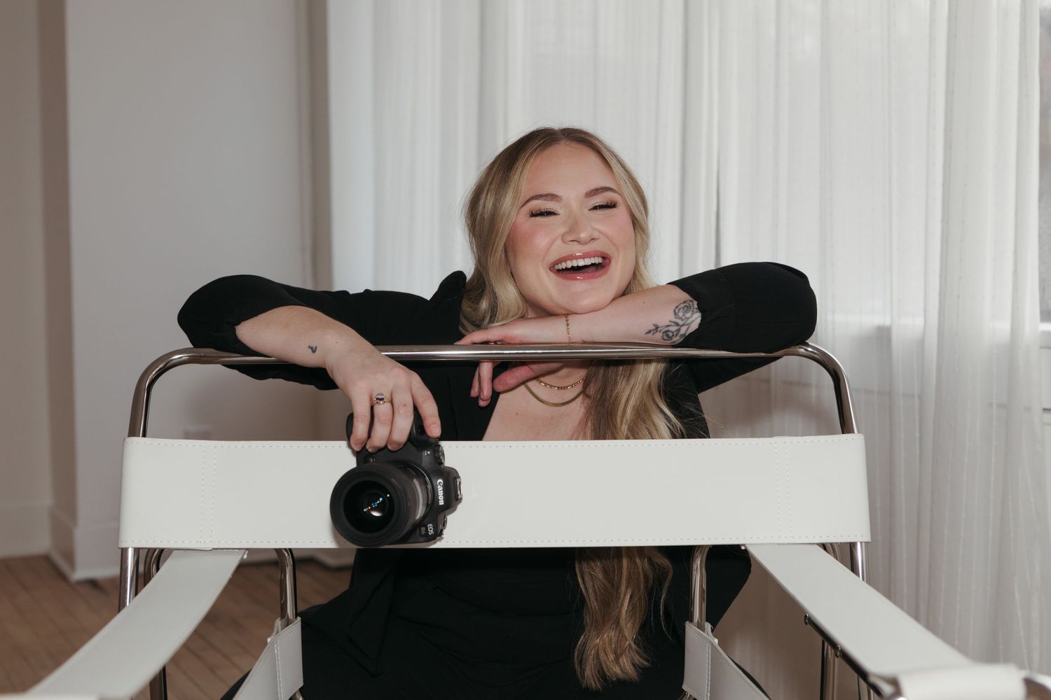 A person laughs joyfully while sitting at a desk in front of a white curtained window.