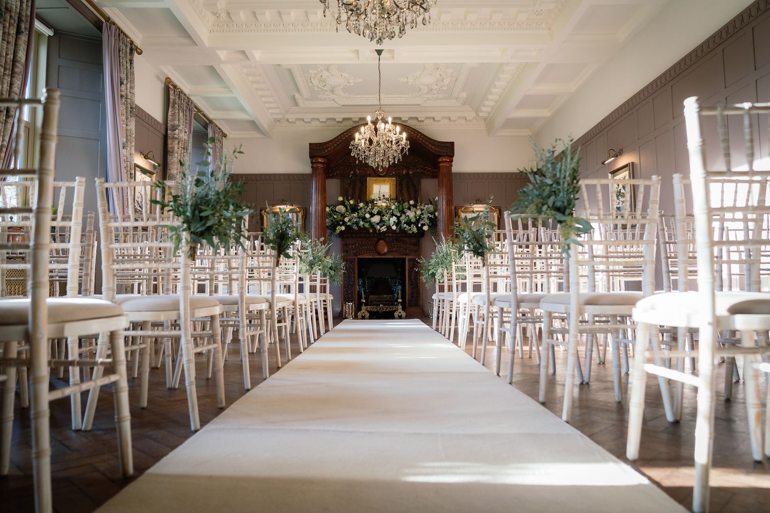 Spacious wedding venue with white chiavari chairs arranged in rows and crystal chandeliers overhead. Ellingham Hall