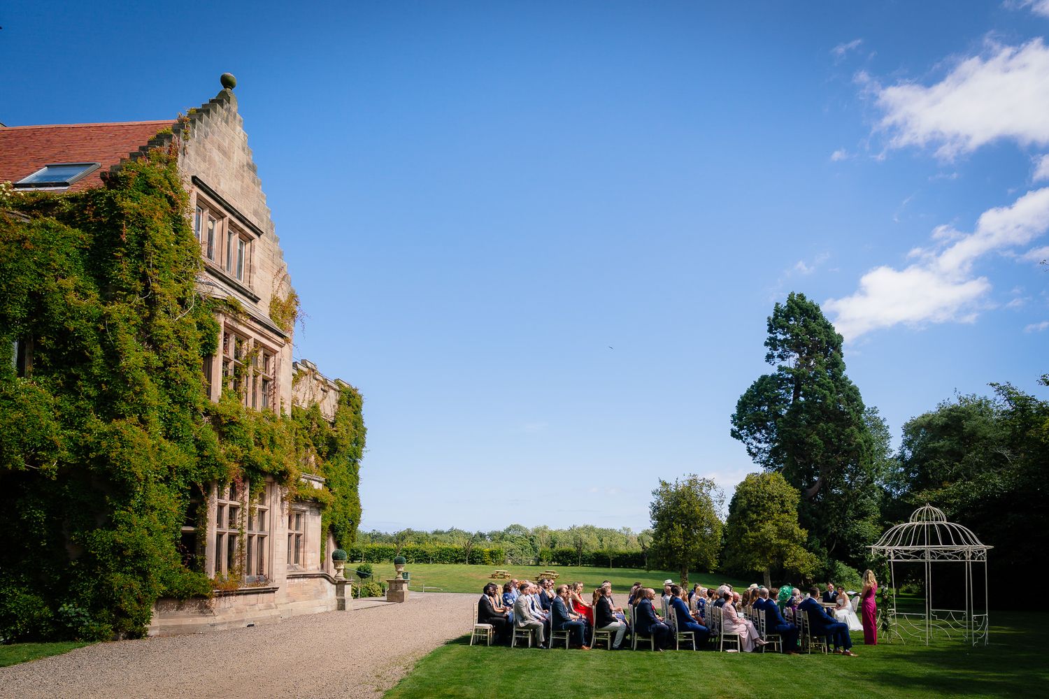 Historic manor house with manicured grounds hosting an outdoor wedding ceremony. Ellingham Hall