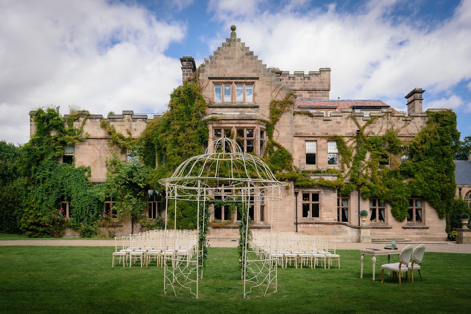 Sweeping view of a stone manor wedding venue with outdoor ceremony setup and metal archways. Ellingham Hall