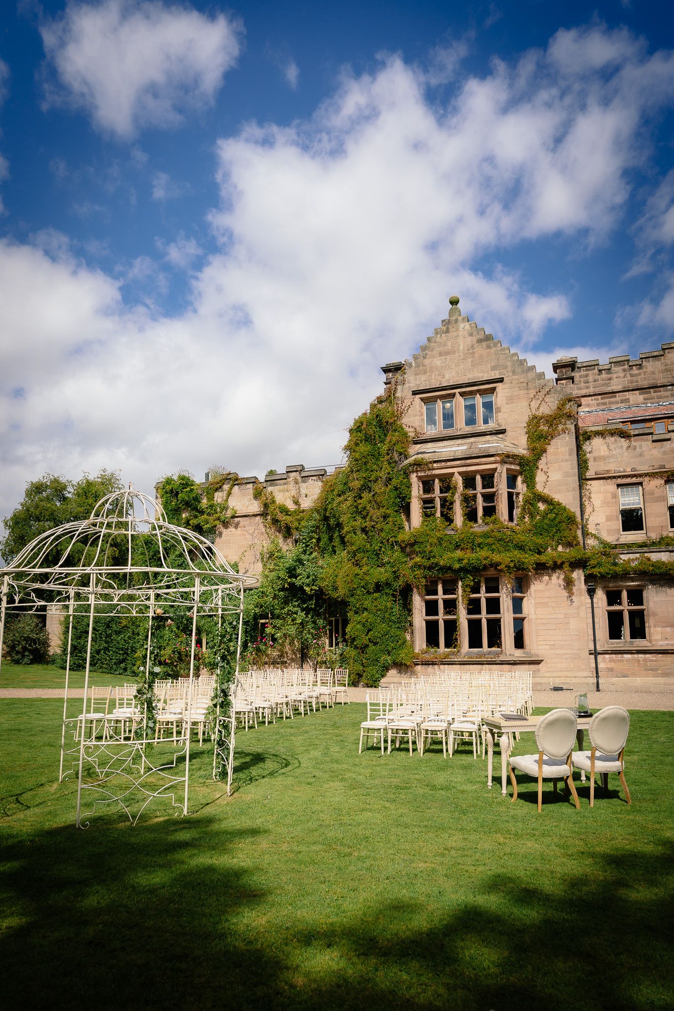 Stone manor house with outdoor wedding ceremony setup featuring white chairs and a metal gazebo. Ellingham Hall