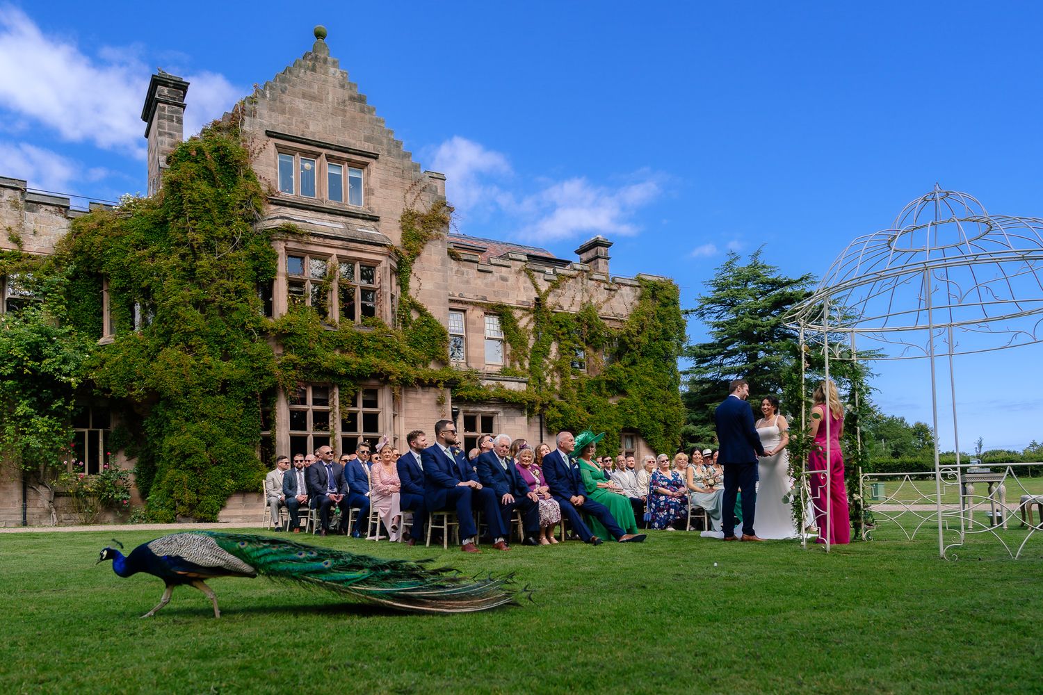 Outdoor wedding ceremony in progress at an elegant stone manor with guests seated on the lawn. Ellingham Hall