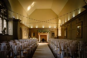 Grand ballroom set up for a wedding with chandeliers and rows of chairs under ambient lighting. Ellingham Hall