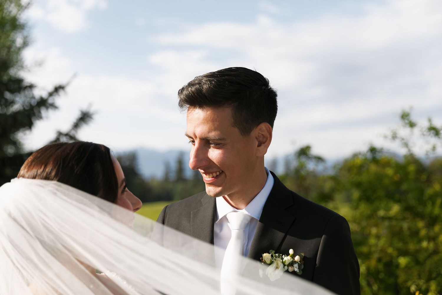 Groom gazing lovingly at bride with flowing veil against a scenic mountain backdrop.